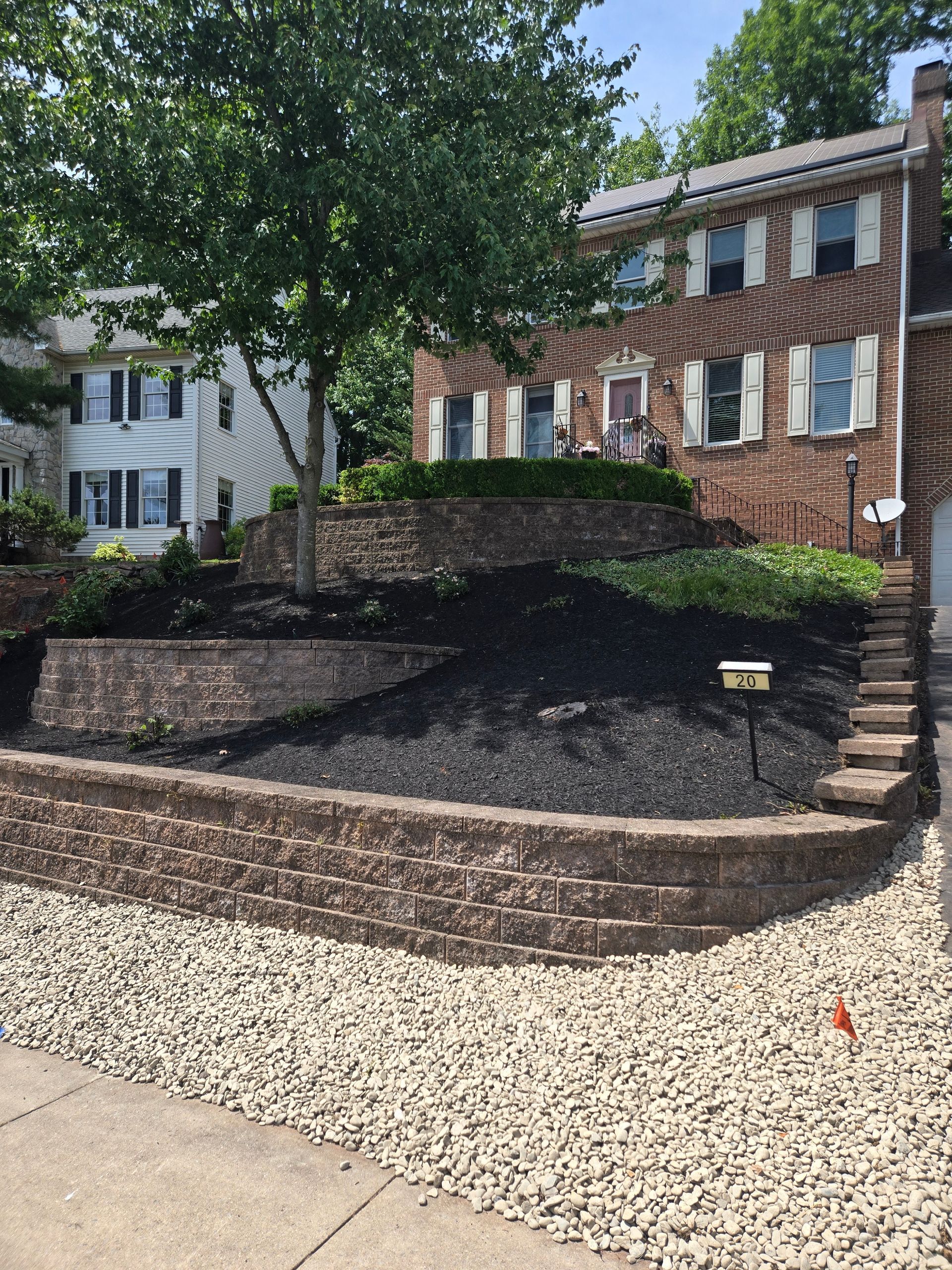 Stone retaining walls and steps front a brick building with white shutters and a large tree.