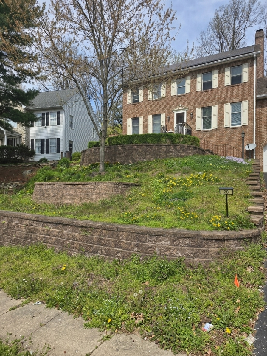 Brick apartment building with terraced stone retaining walls, grassy slope, and neighboring houses under cloudy sky