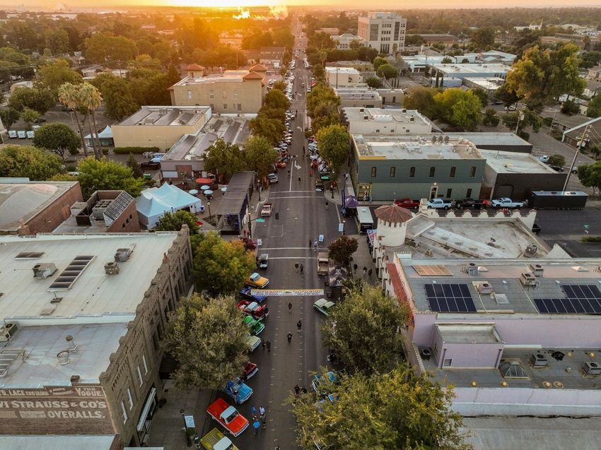 Aerial view of a street lined with trees and buildings at sunset, with cars and pedestrians below.