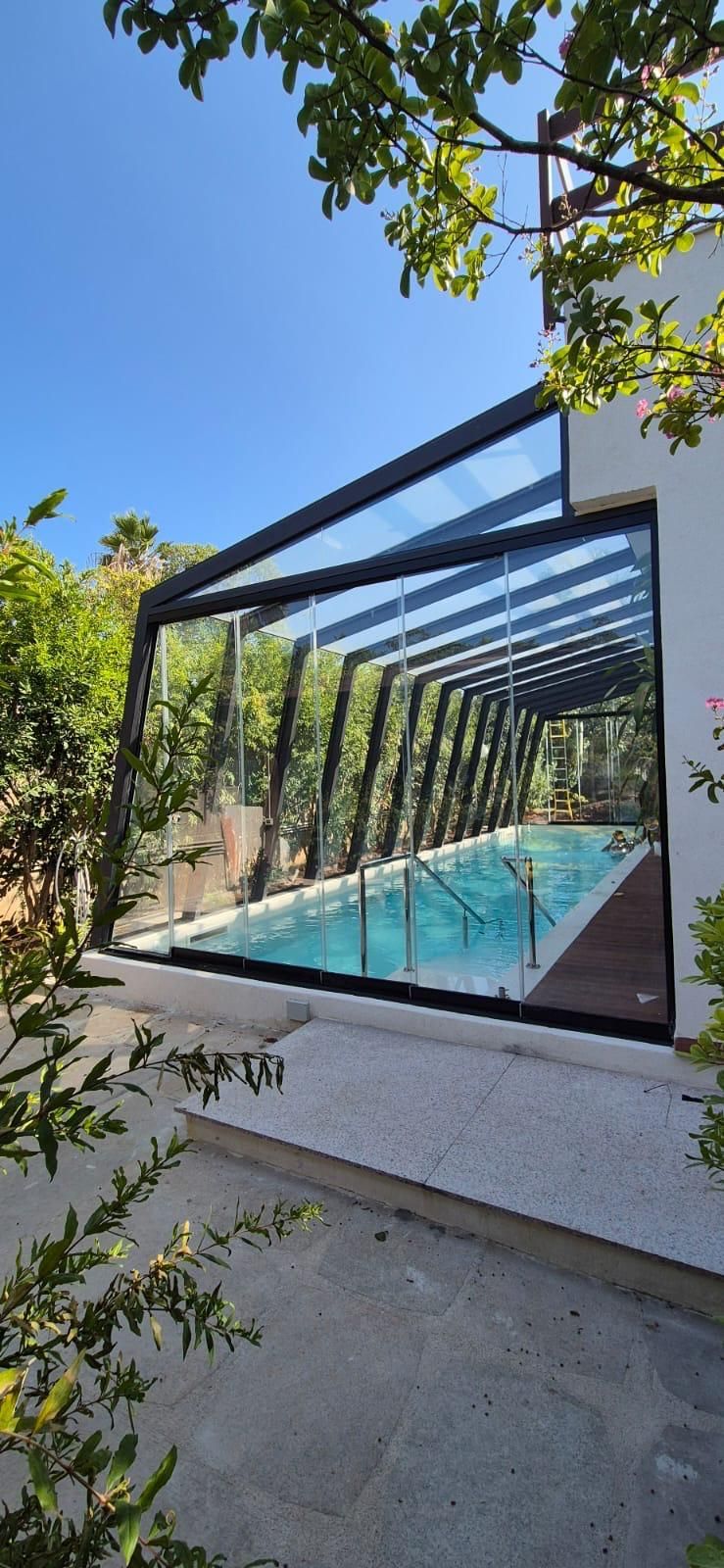 Una piscina coperta con pareti in vetro. Alberi intorno alla piscina. Il cielo è azzurro.