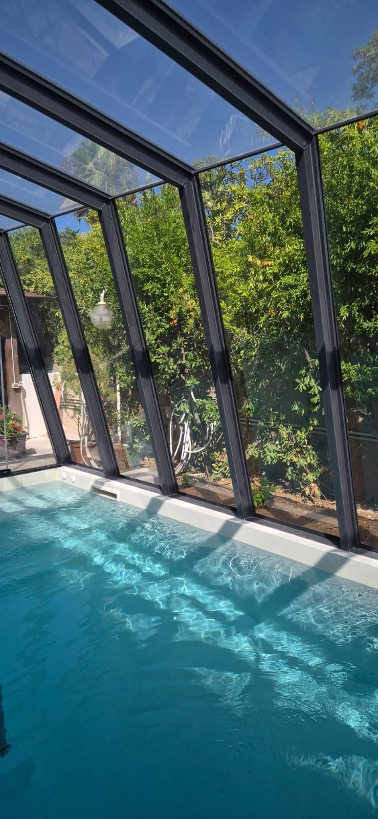 Una piscina coperta con tetto e pareti in vetro trasparente, acqua azzurra e vista sugli alberi verdi.