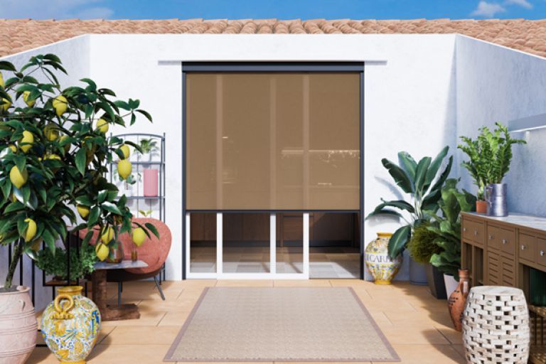 Patio esterno con tenda da sole marrone, piante e un tappeto intrecciato.