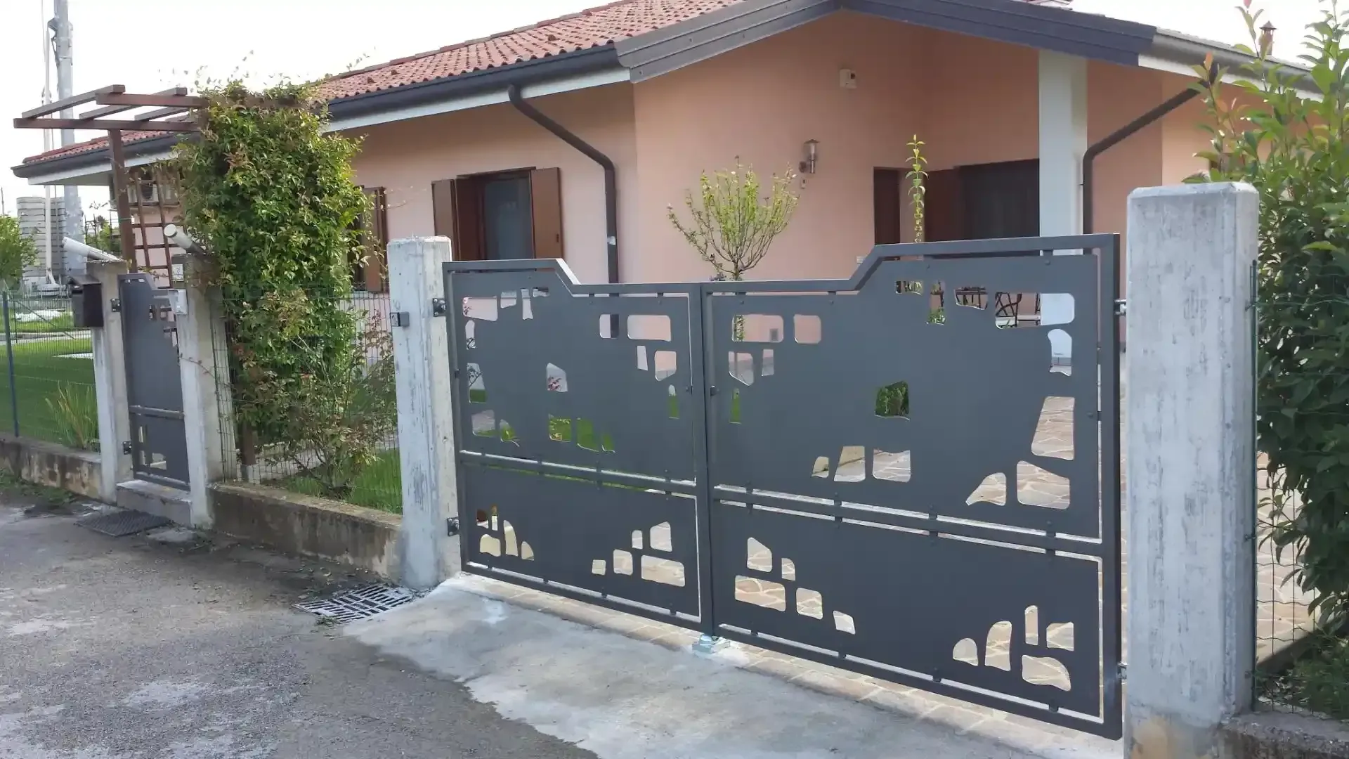 Cancello in metallo grigio scuro, pali in cemento, piccola casa beige con tetto e persiane marroni.