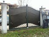 Cancello in metallo scuro tra pilastri in cemento con volute decorative.