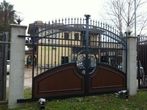 Cancello in ferro decorato marrone e nero con un edificio sullo sfondo. È fiancheggiato da due pilastri grigi con luci.