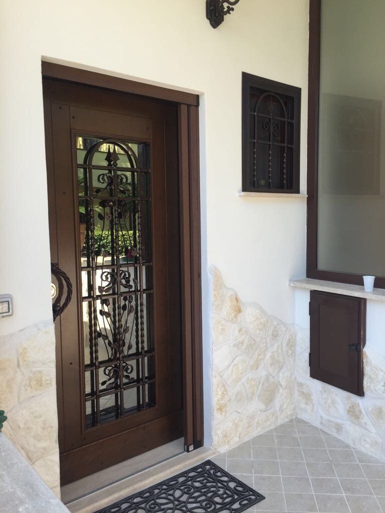 Porta d'ingresso in legno con elementi decorativi in ferro battuto, accanto a una piccola finestra e a una sezione di muro in pietra.