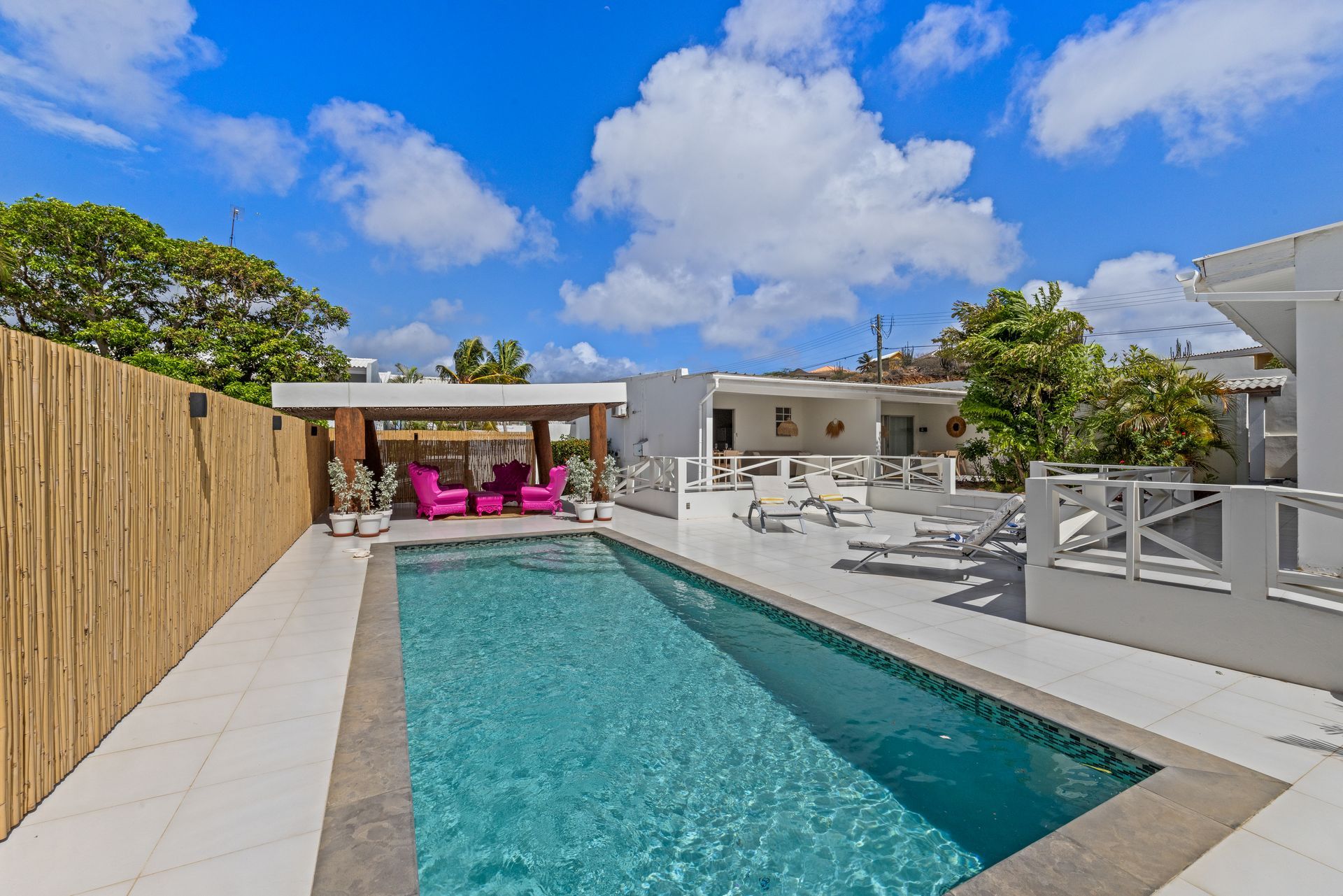 Swimming pool in front of white building with outdoor seating and palm trees under a partly cloudy sky.