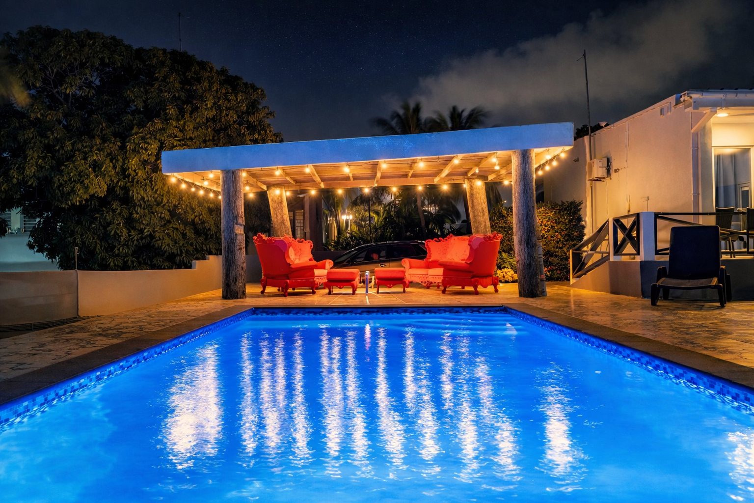Poolside lounge area at night; illuminated pool, red furniture under pergola, string lights.