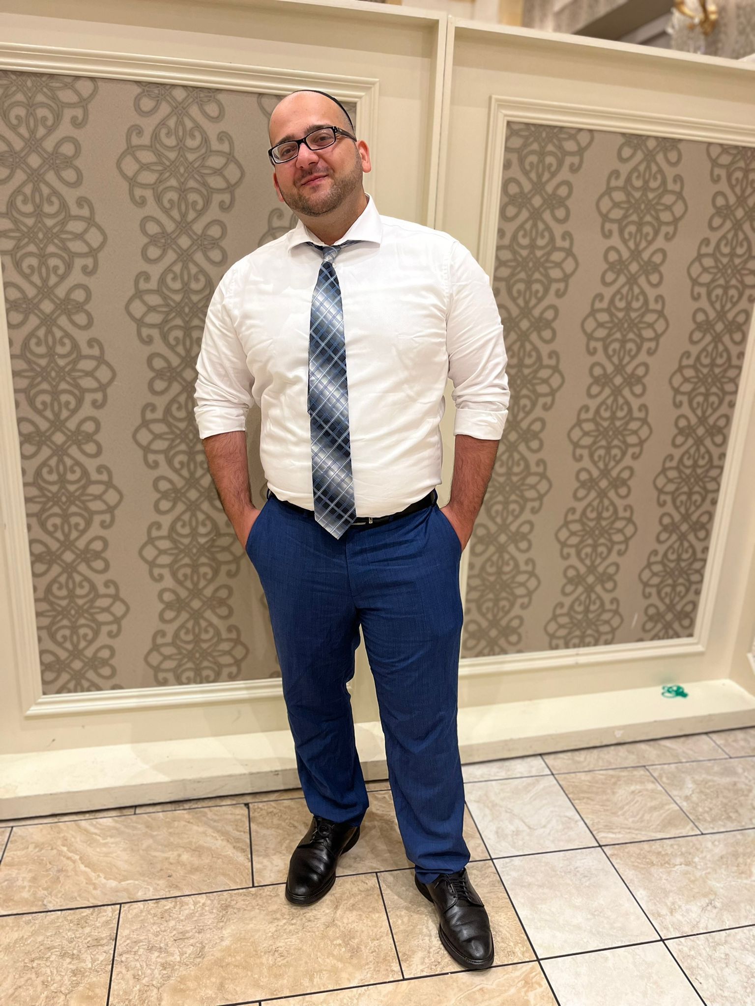Man in white shirt and blue tie standing indoors against patterned wall panels.