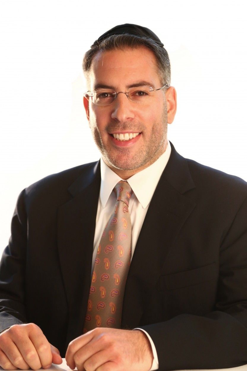 Man in a dark suit and patterned tie smiling at a desk against a white background