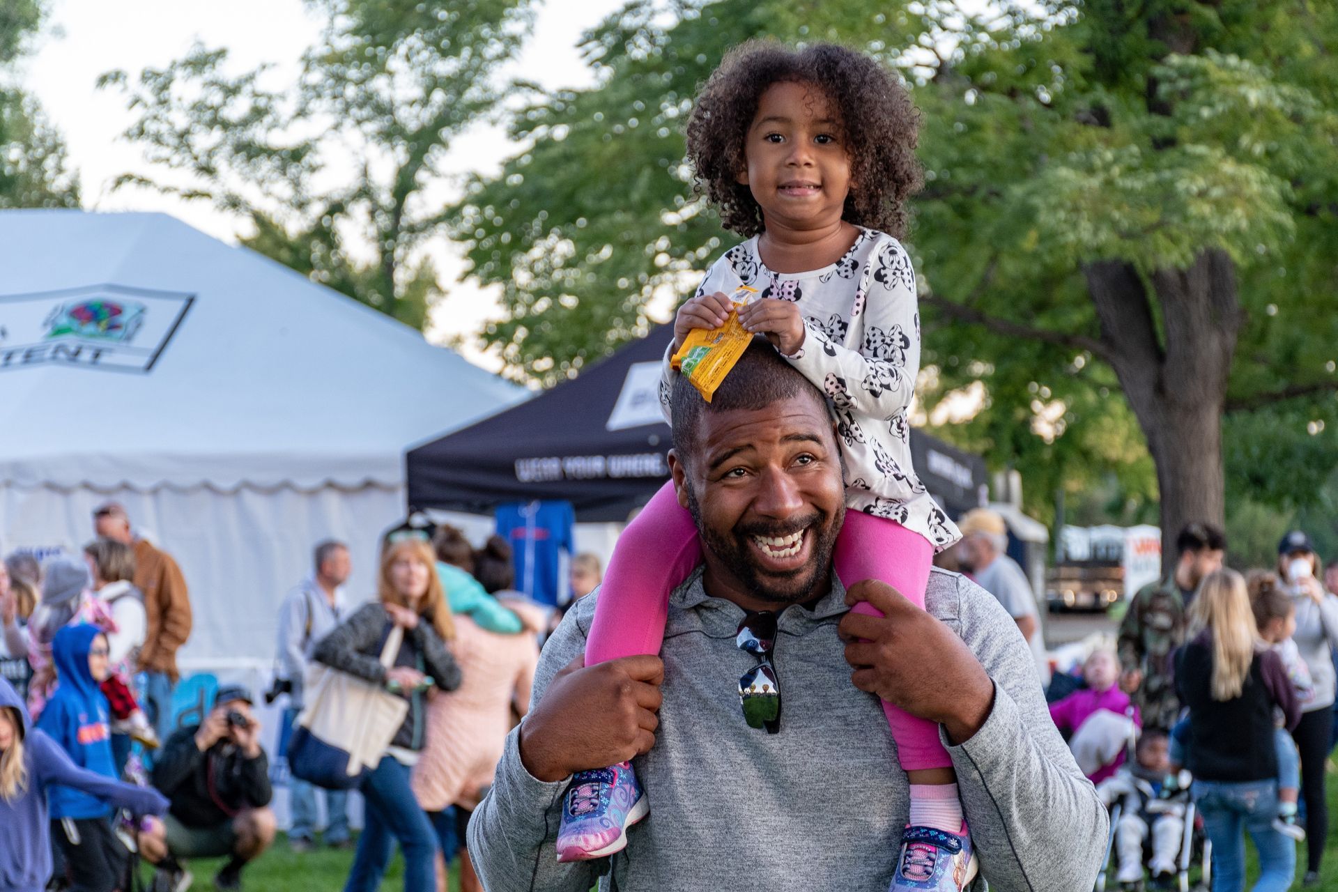 Man smiling with child on his shoulders at an outdoor community event with tents and crowd