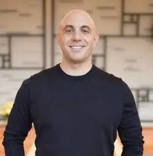 Smiling bald man in a dark sweater standing indoors against a blurred patterned wall