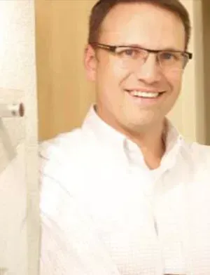Smiling man in a white shirt and glasses standing indoors by a doorway