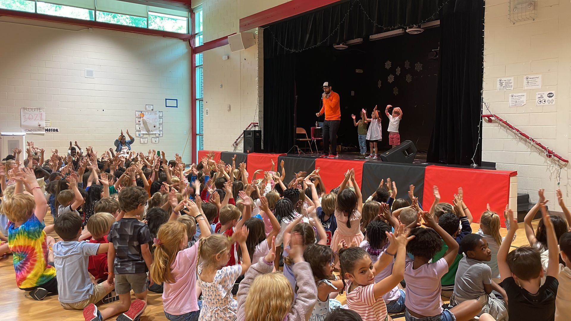 Audience of children clapping at a stage performance in a gymnasium, with performers onstage.