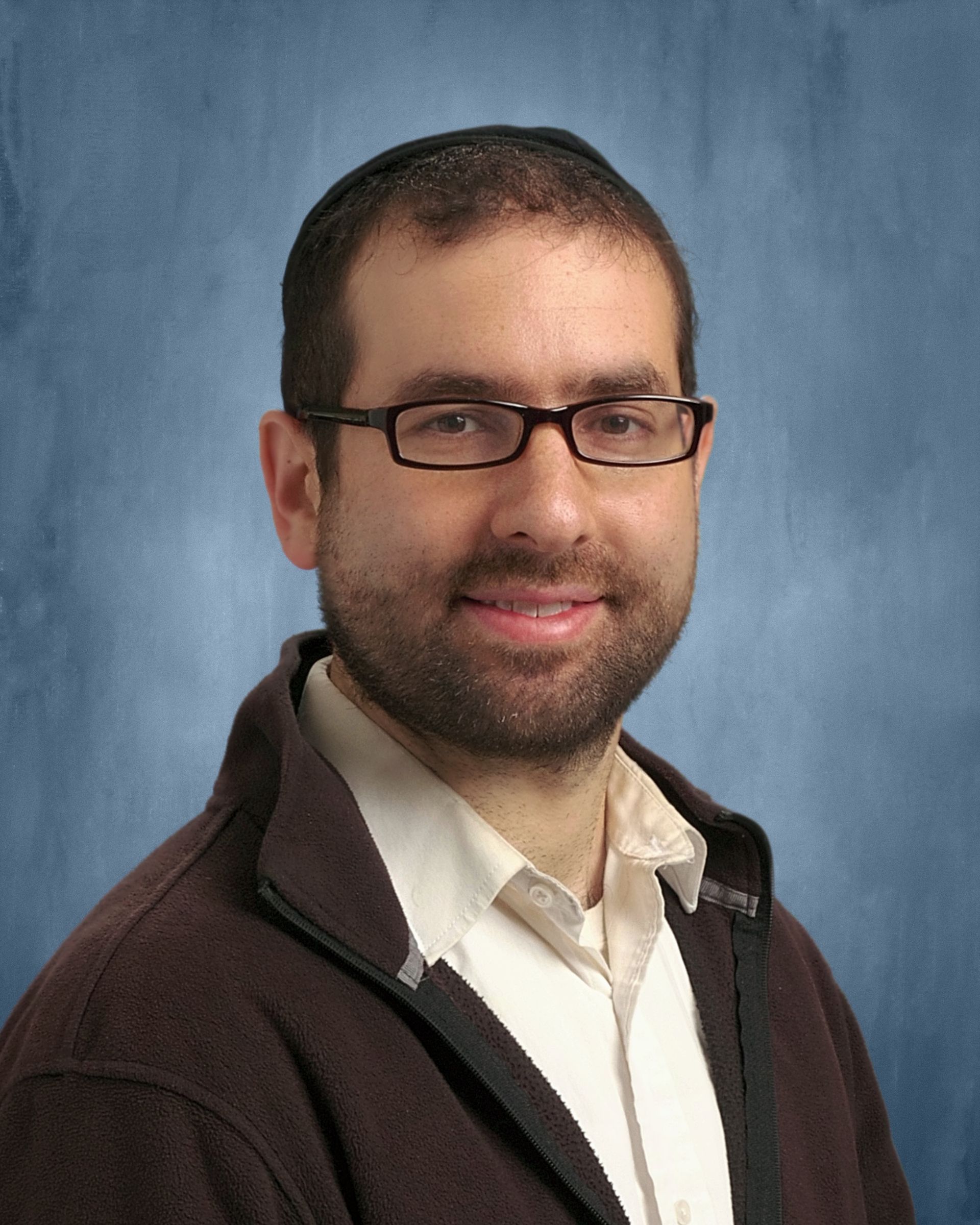 Smiling man with glasses and short beard in a white shirt and dark jacket against a blue backdrop