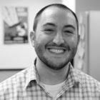 Smiling man in a checkered shirt, photographed in grayscale against a blurred indoor background