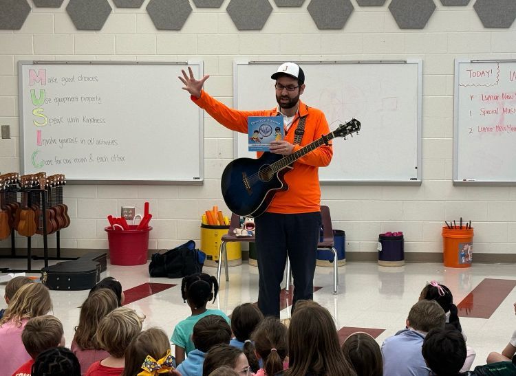 Teacher with guitar leading a seated classroom lesson, waving in front of a whiteboard.