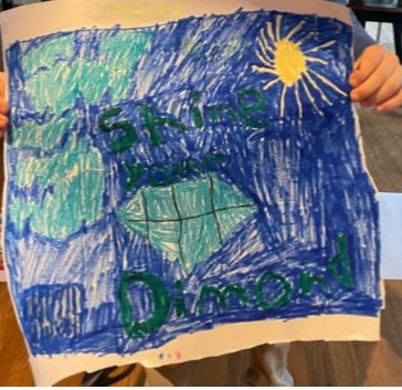 Child holding a blue crayon drawing of a green diamond-shaped logo with a yellow sun in the corner