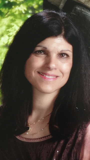Smiling woman with long dark hair, wearing a burgundy top, posed outdoors near greenery