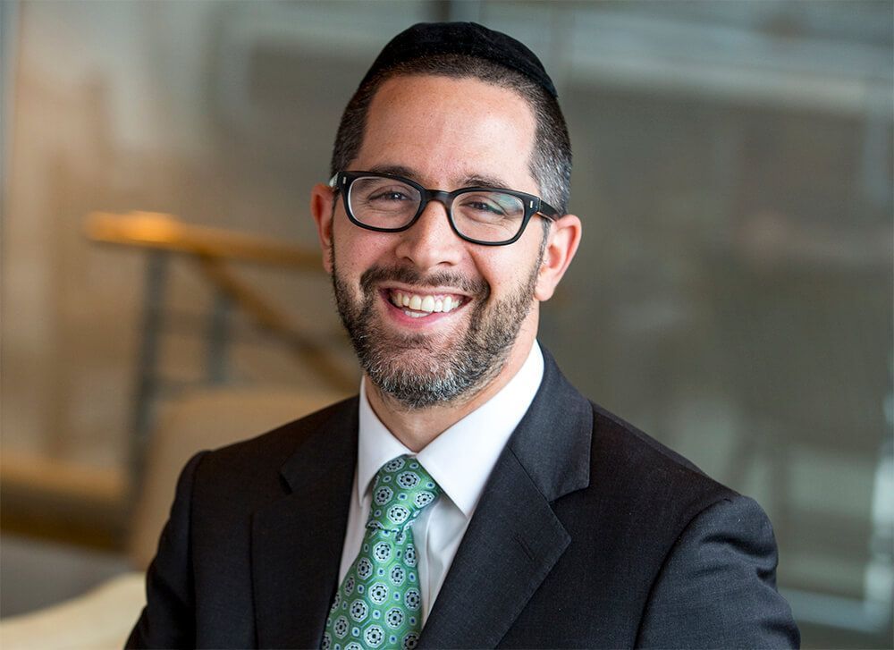 Smiling man in glasses and suit with green tie, standing indoors against a blurred background