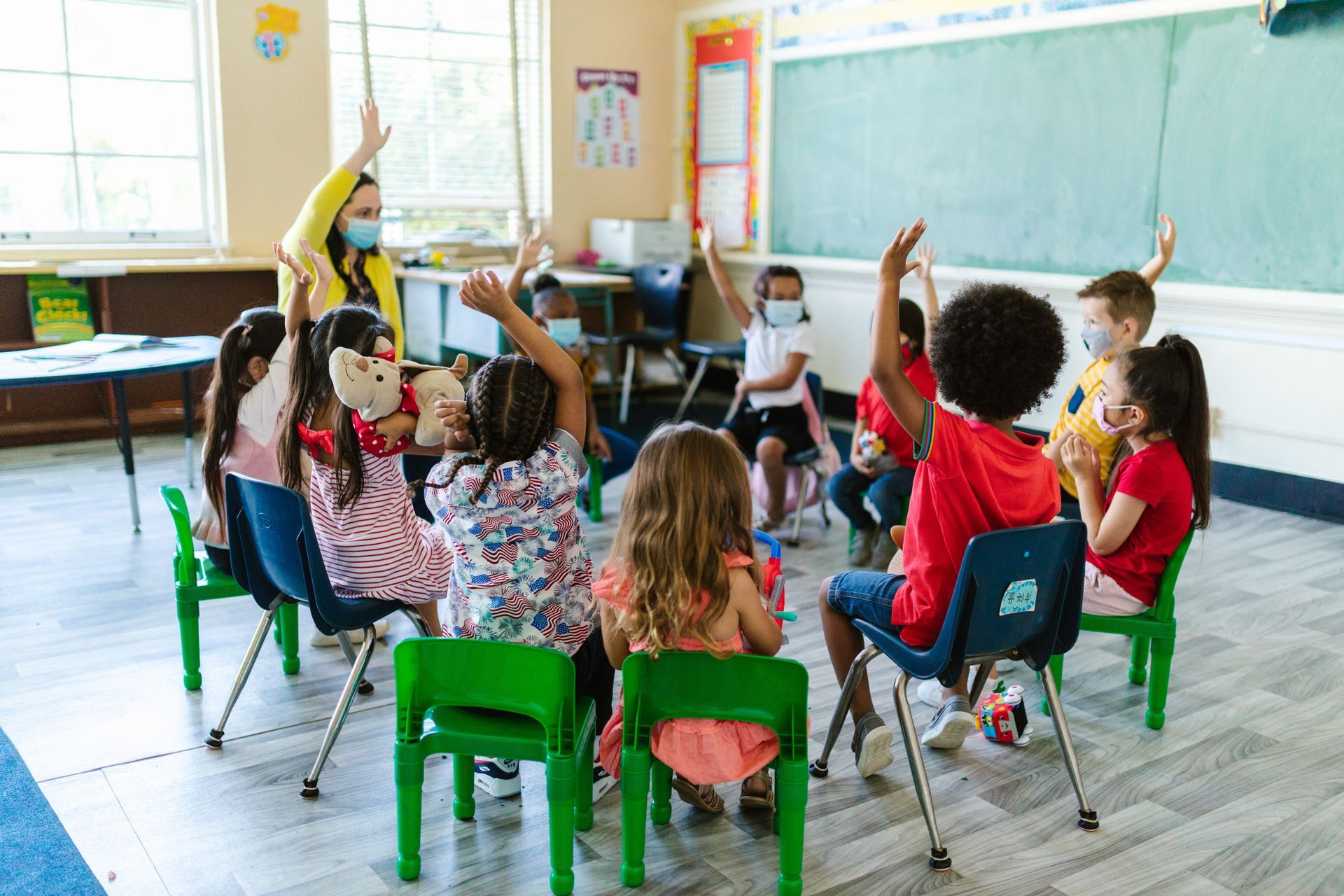 Children raising hands in a classroom circle with a teacher near a chalkboard