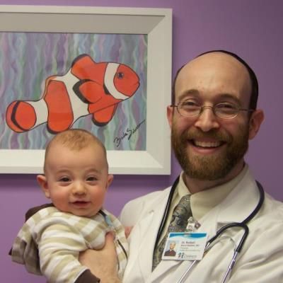 Smiling man holding a baby in a doctor’s office, with a clownfish painting on the wall.