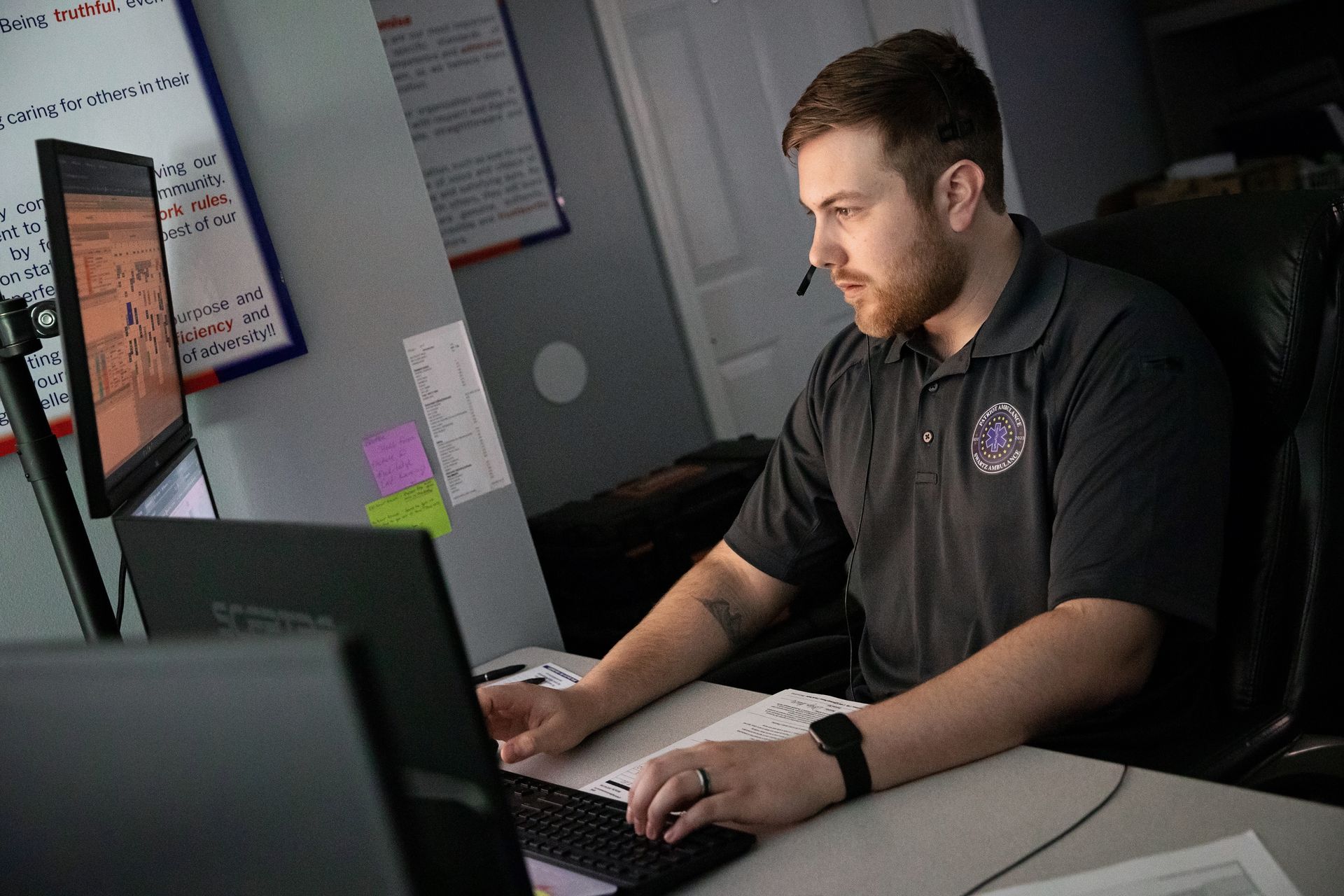 Man in headset at desk using a laptop with two monitors, in office setting.