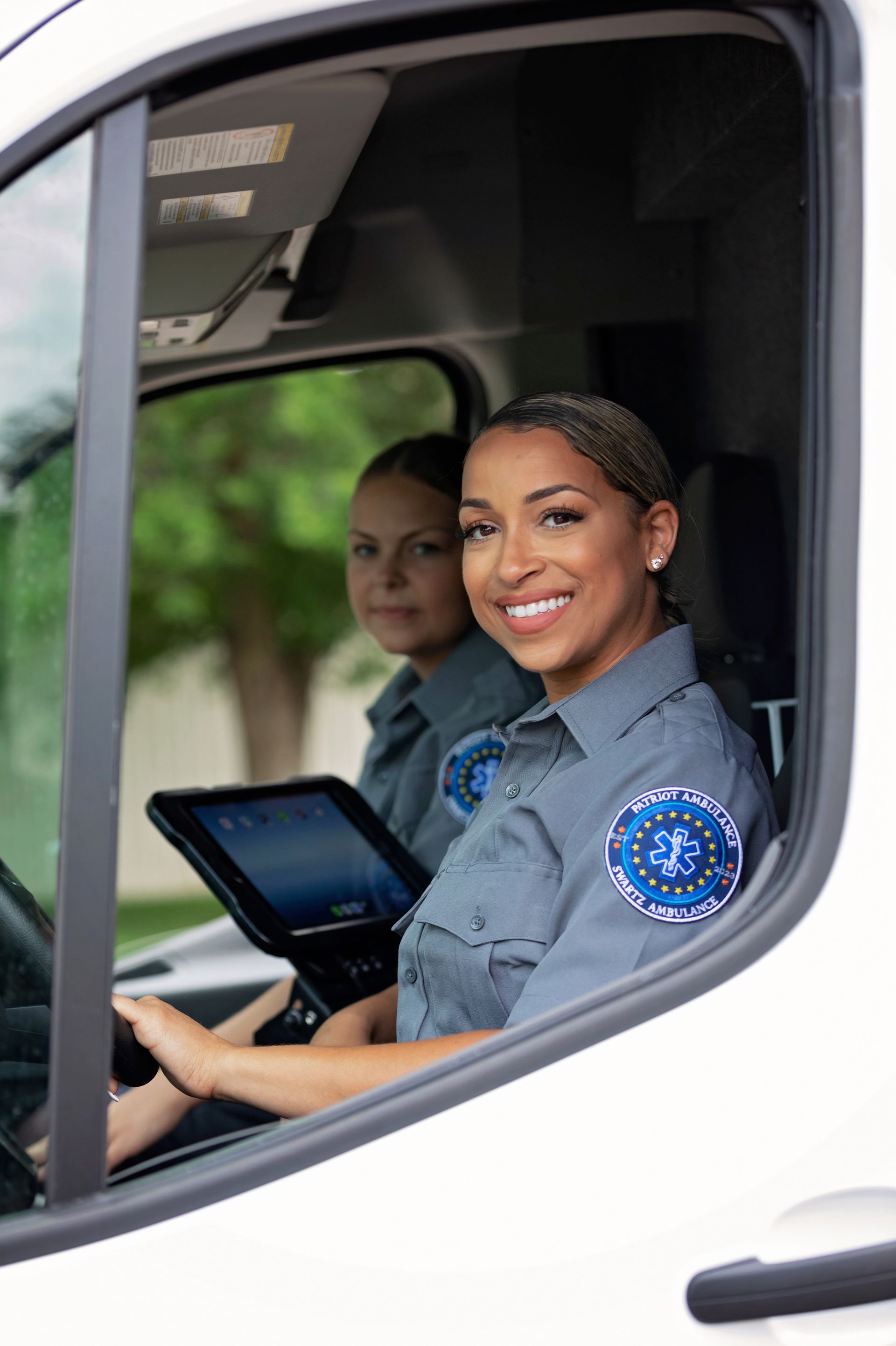 Two people in gray uniforms smiling from an ambulance. One operates a tablet while the other looks on.