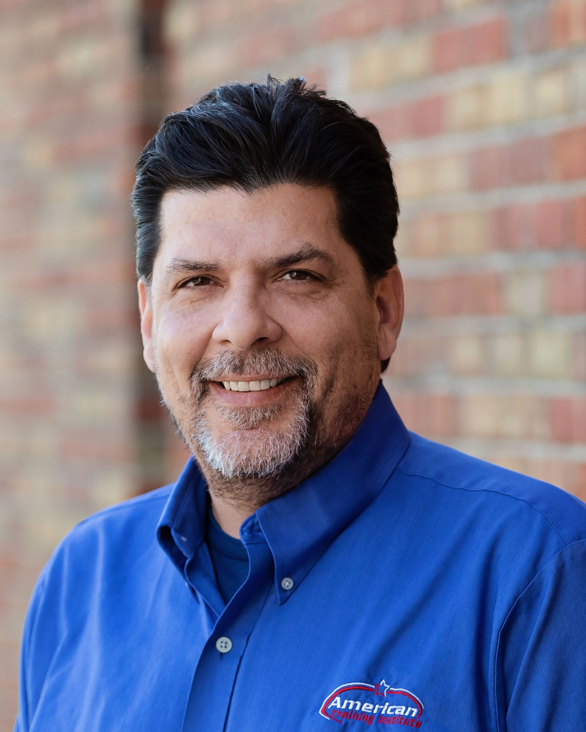 Smiling man in blue shirt in front of a brick wall. The shirt has a logo on it.
