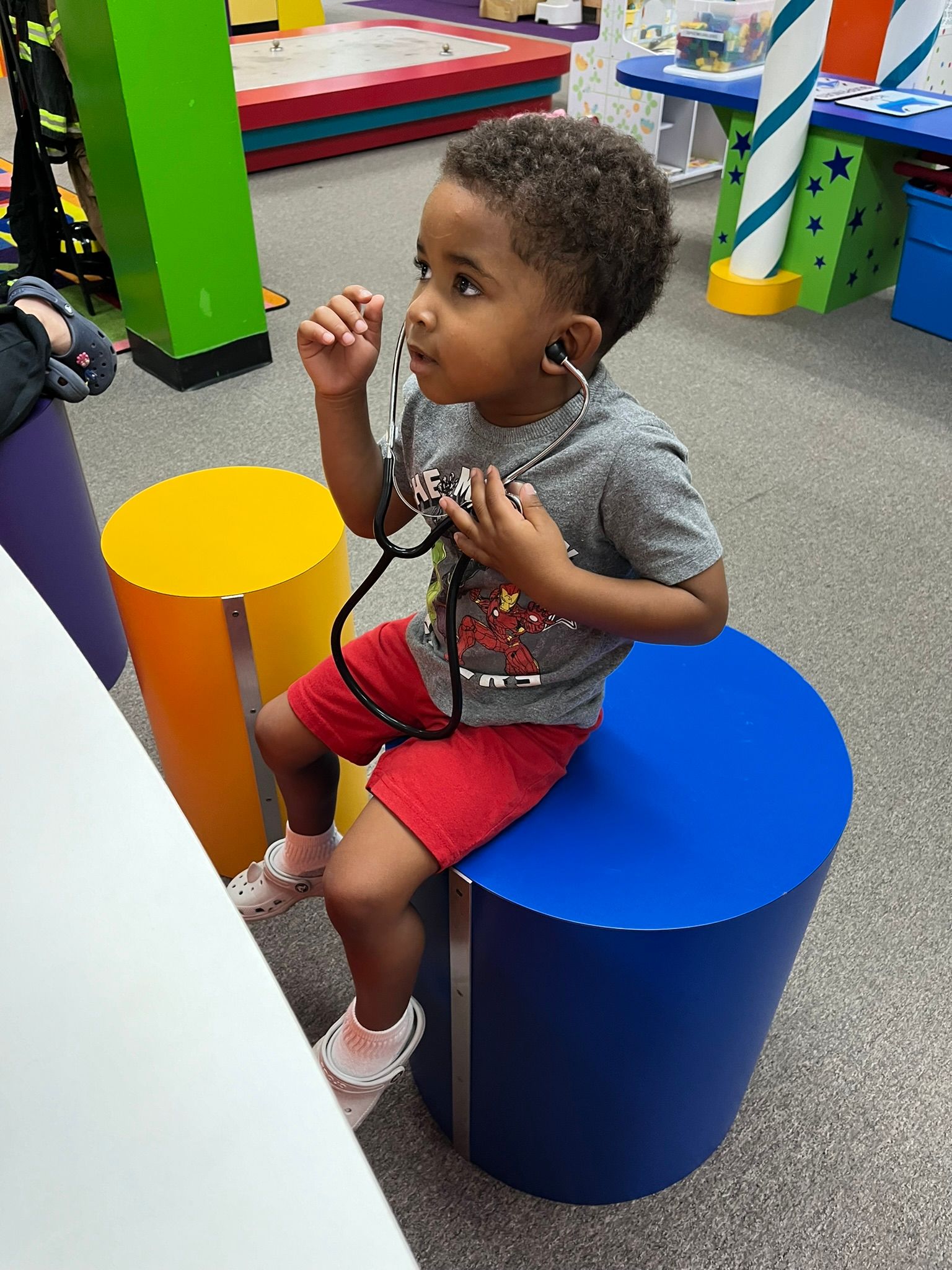 Young child seated, using a stethoscope. Wearing red shorts and a gray shirt. Playroom setting, colorful cylinders.