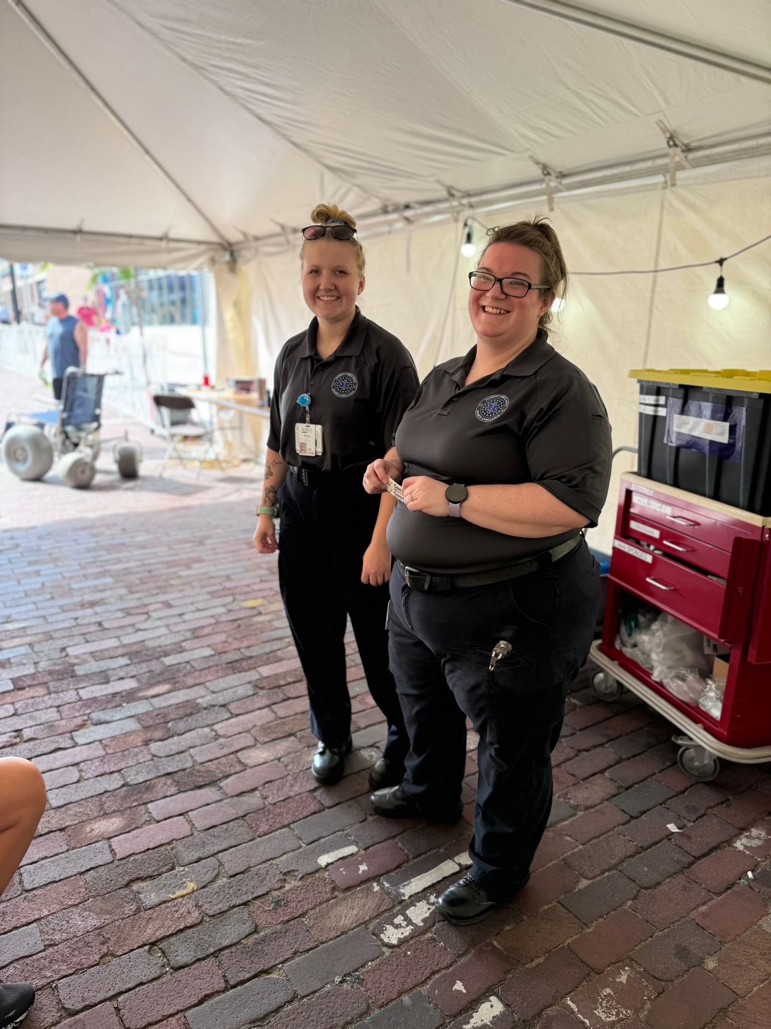 Two uniformed EMTs smile under a tent; one with a high bun, one with glasses, near equipment.