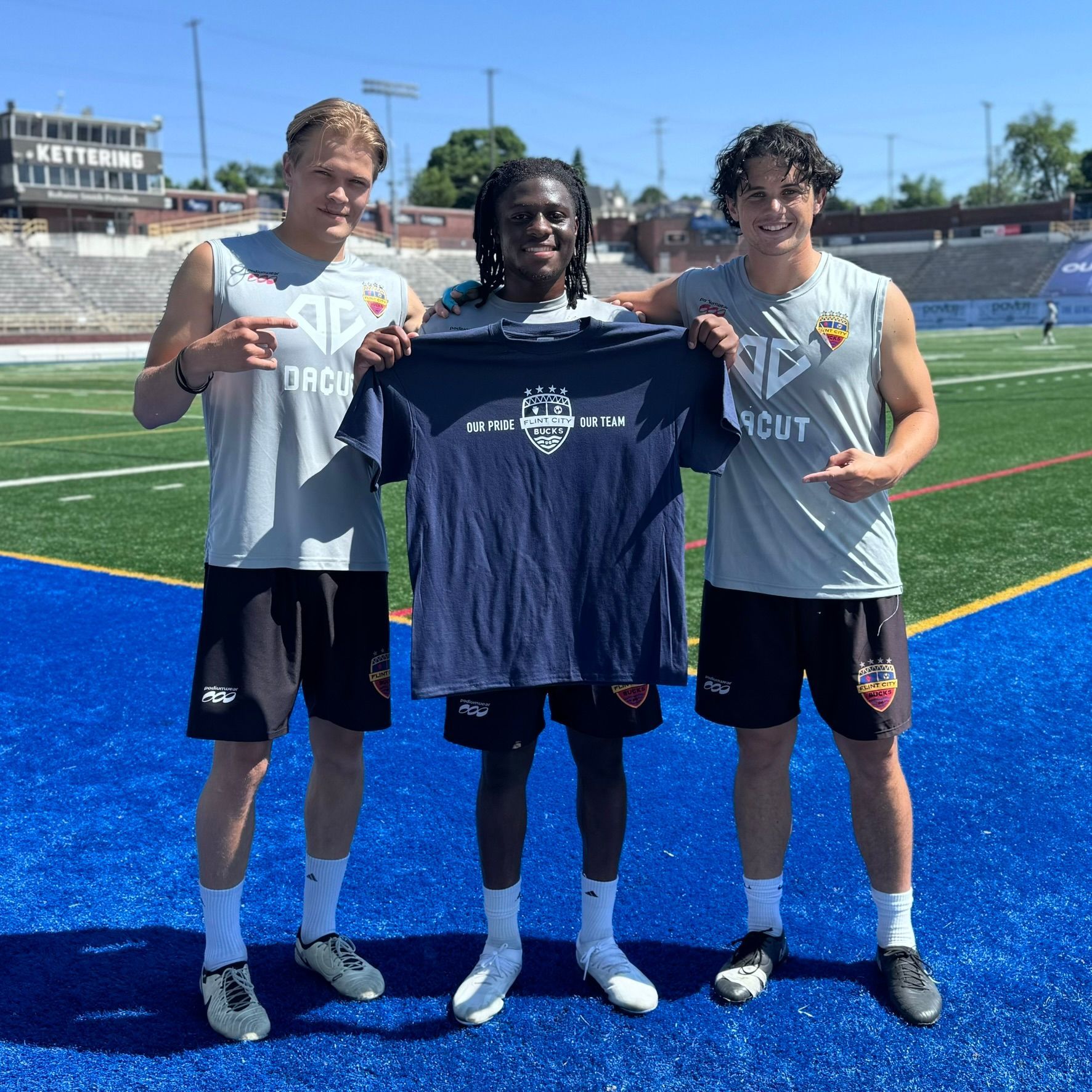 Three athletes on a blue field holding a dark blue shirt; two pointing at the shirt.