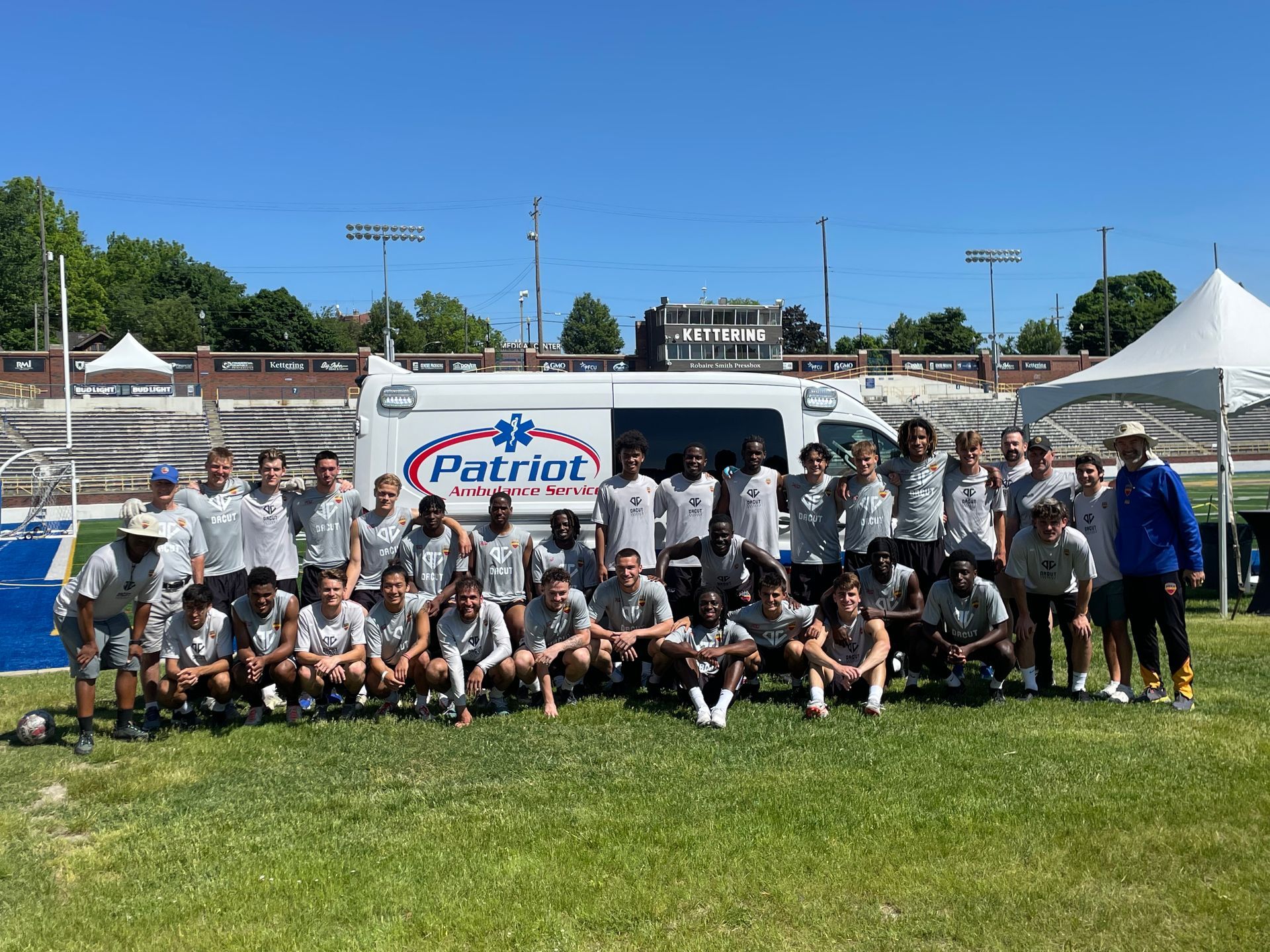 Group of people posing in front of an ambulance on a grassy field; blue sky, stadium in background.