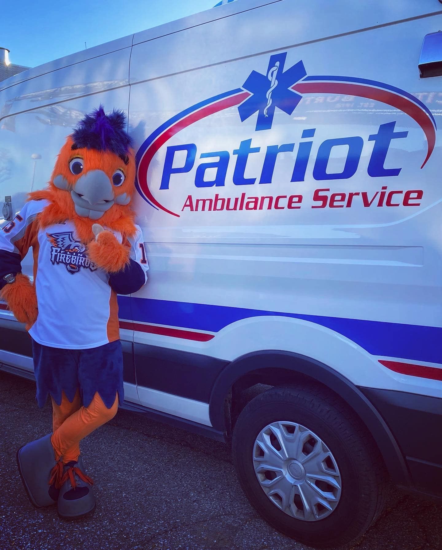 Mascot with orange and purple feathers, leaning on a Patriot Ambulance Service vehicle.