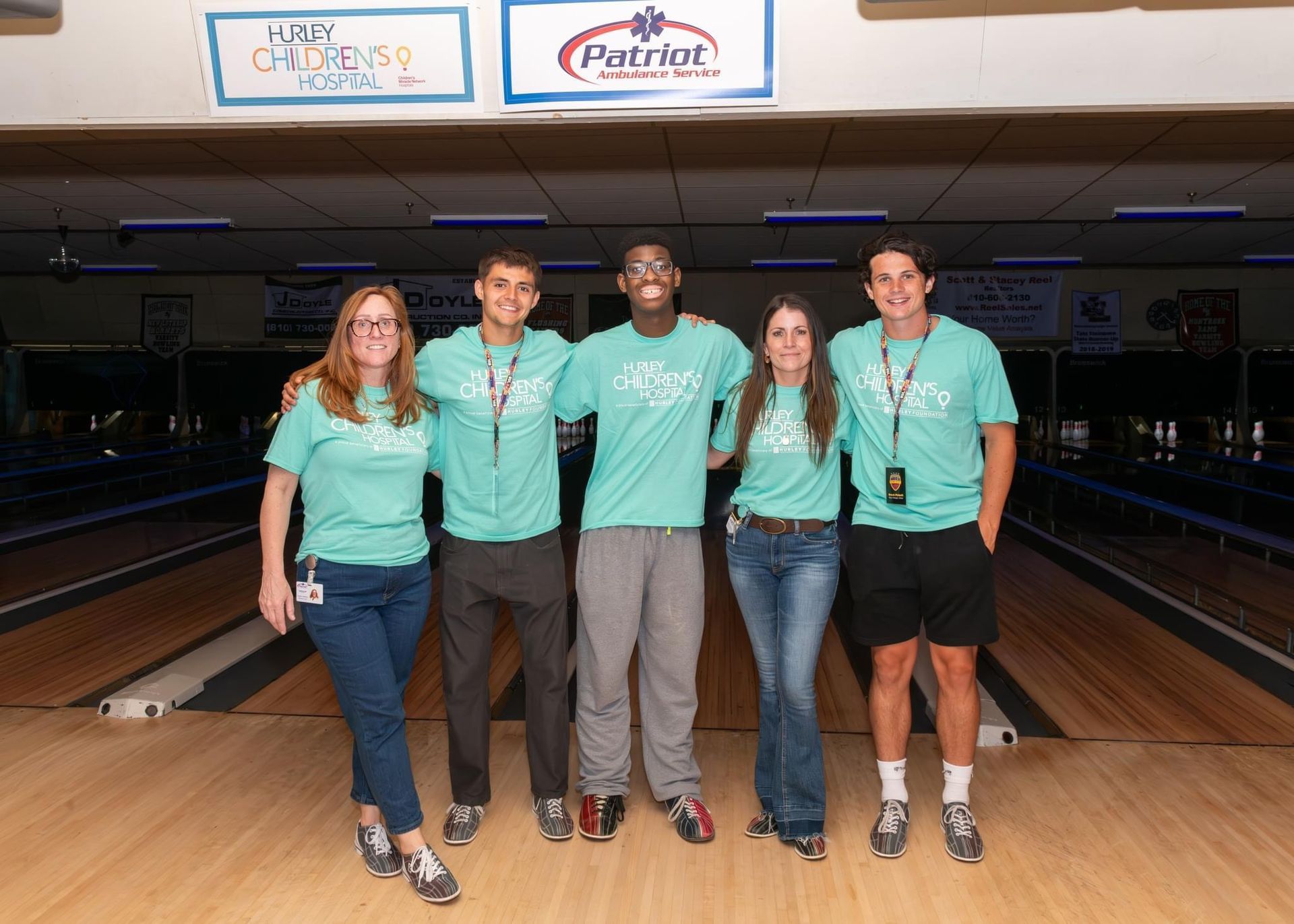 Five people in matching teal shirts stand in a bowling alley, smiling.