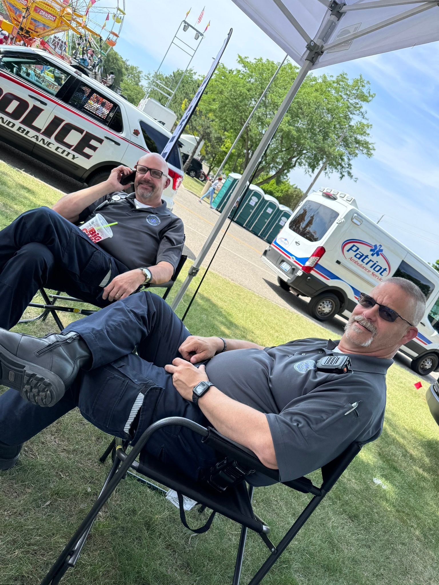 Two police officers relaxing in chairs under a canopy at an outdoor event, with police vehicles visible.
