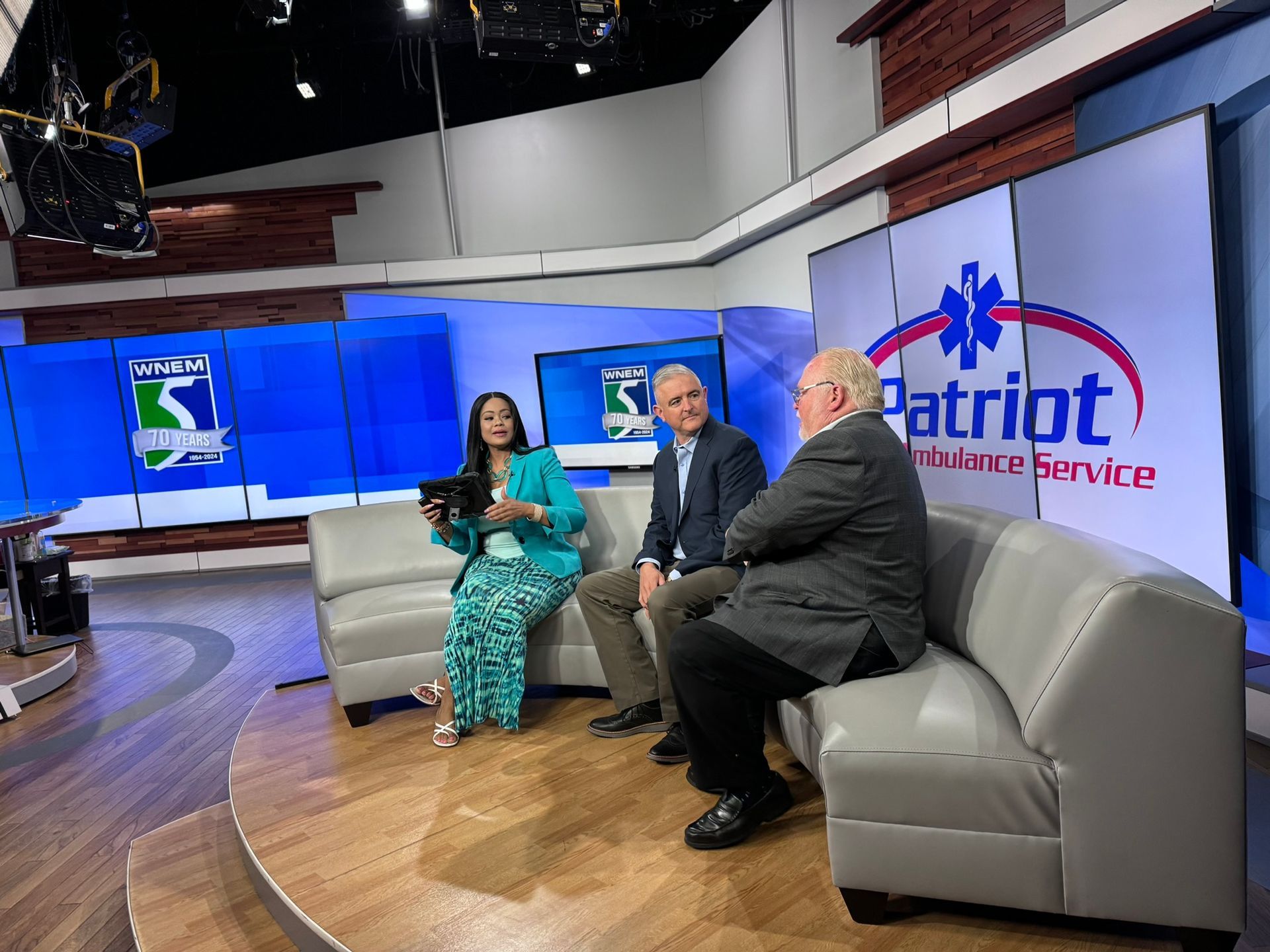 Three people on a TV set; woman in teal, two men in suits, blue backdrop with logo, grey couch.