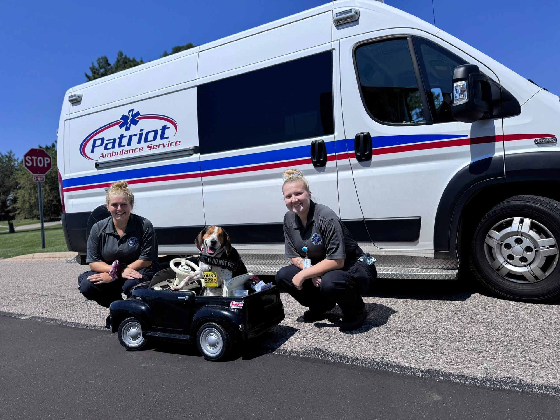 Two women and a dog pose with a toy car next to a Patriot medical transport van.