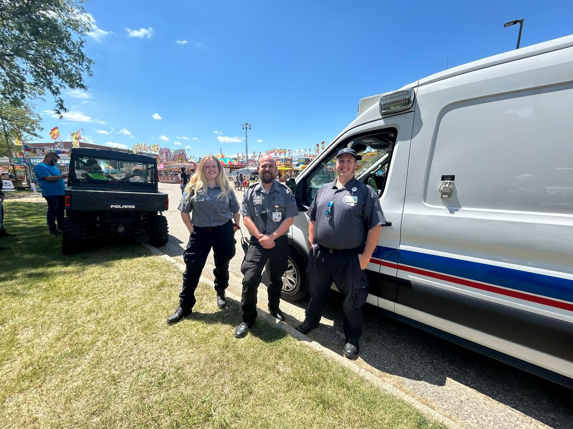 Three people in uniform stand near an ambulance and an ATV on a grassy field under a blue sky.