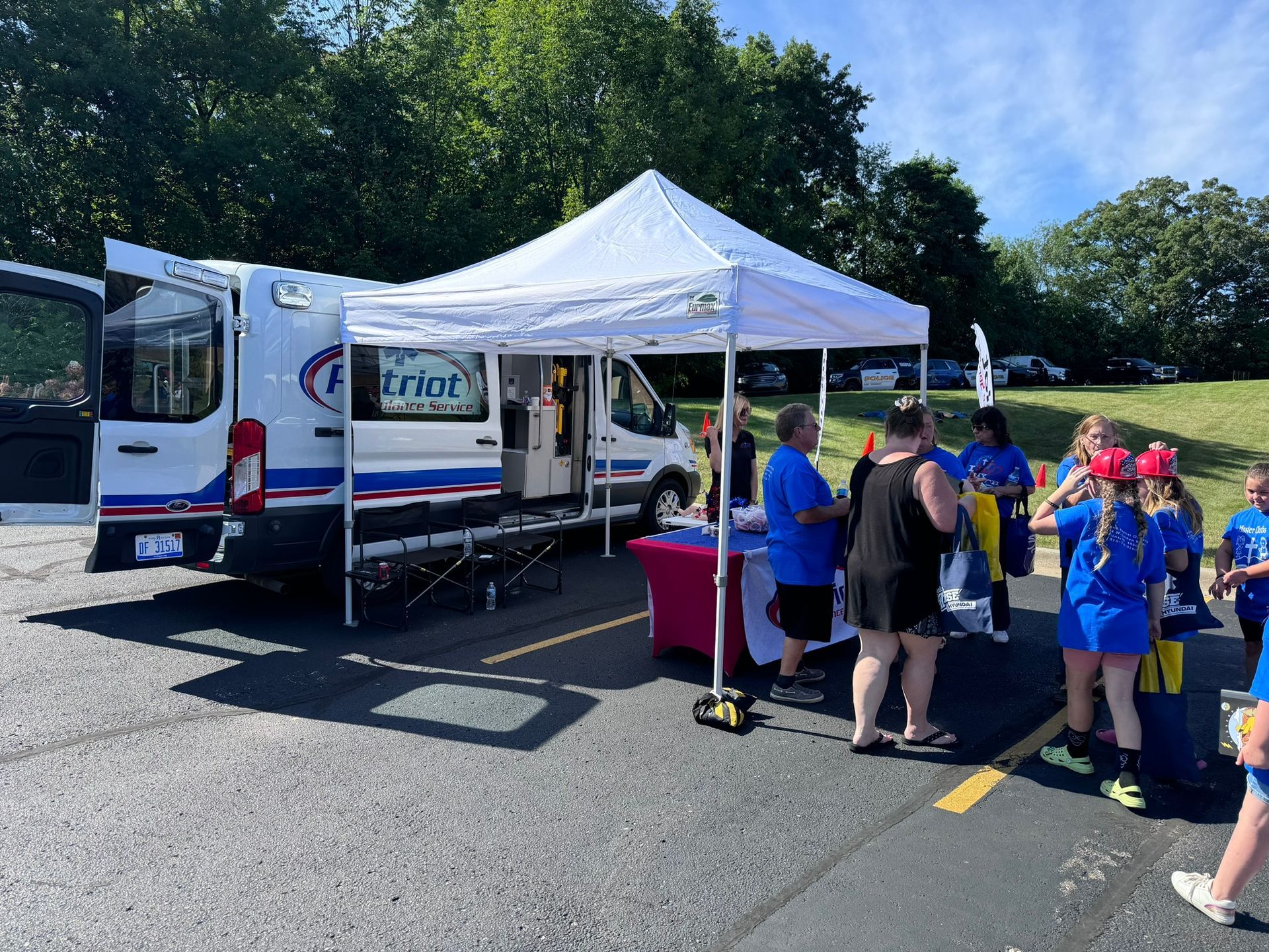 Event with a white tent, parked van, and people in a parking lot on a sunny day.