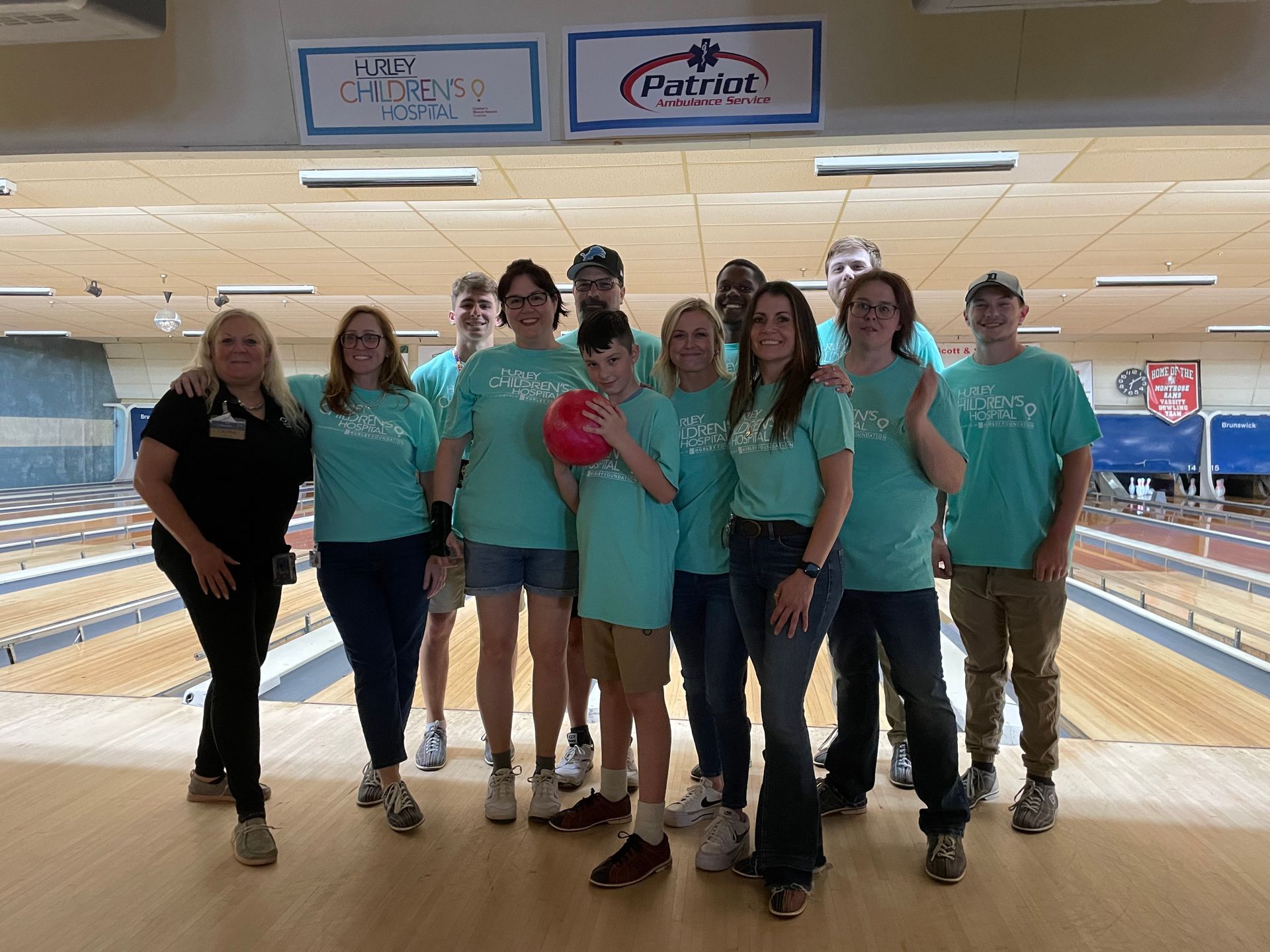 Group in teal shirts at bowling alley, one holding a ball, posing by lanes.