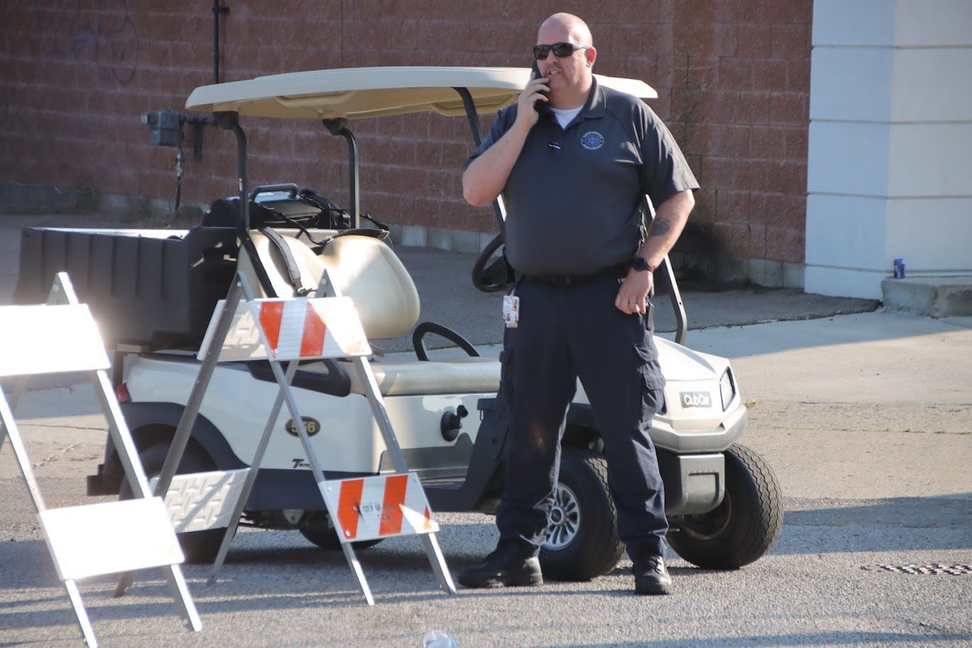 Security guard in dark uniform talks on phone, standing near a golf cart and traffic barricade.
