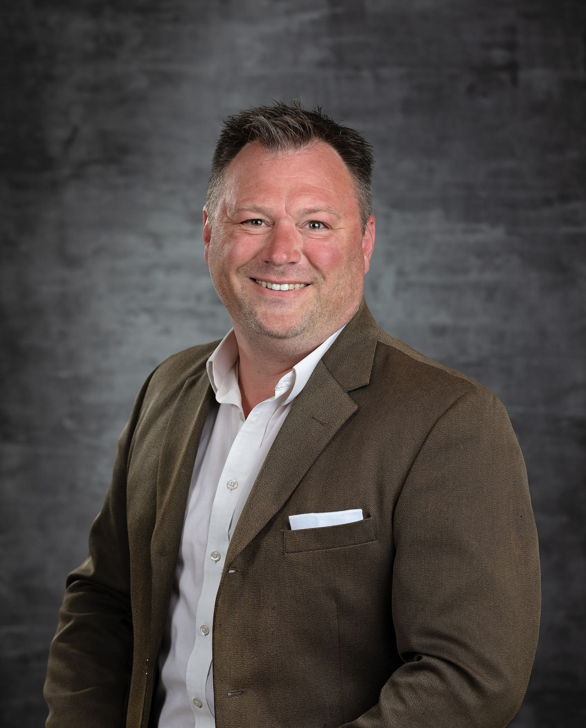Man in a brown patterned blazer smiles; white shirt, gray background.