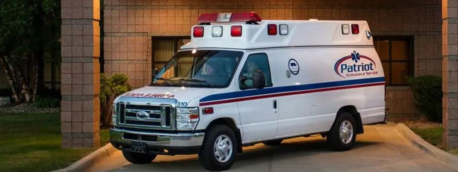 White ambulance parked in front of a building with Patriot Emergency Services logo.