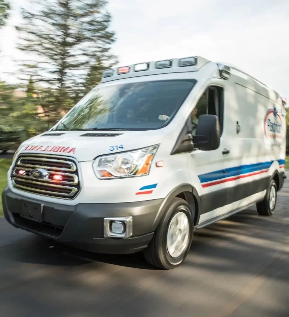 White ambulance with red and blue stripes and flashing lights on a road.