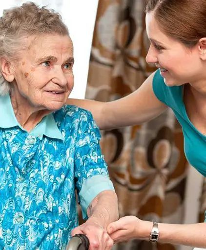 Woman assisting elderly person using a walker, indoors. Blue clothing, supportive arm, smiles.