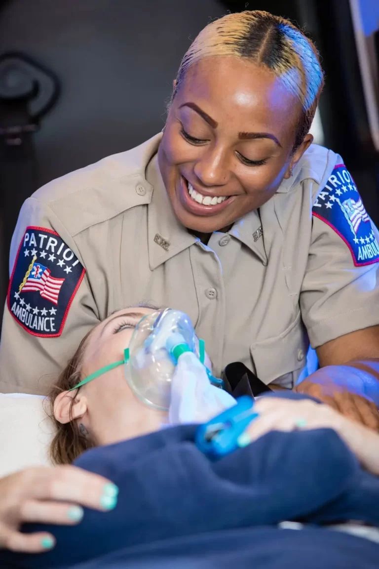 Paramedic in tan uniform smiles at patient wearing oxygen mask inside ambulance.