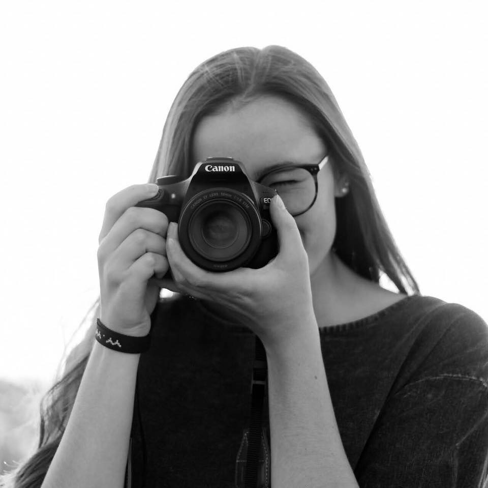 Woman with glasses holding a Canon camera to her eye, outdoors.