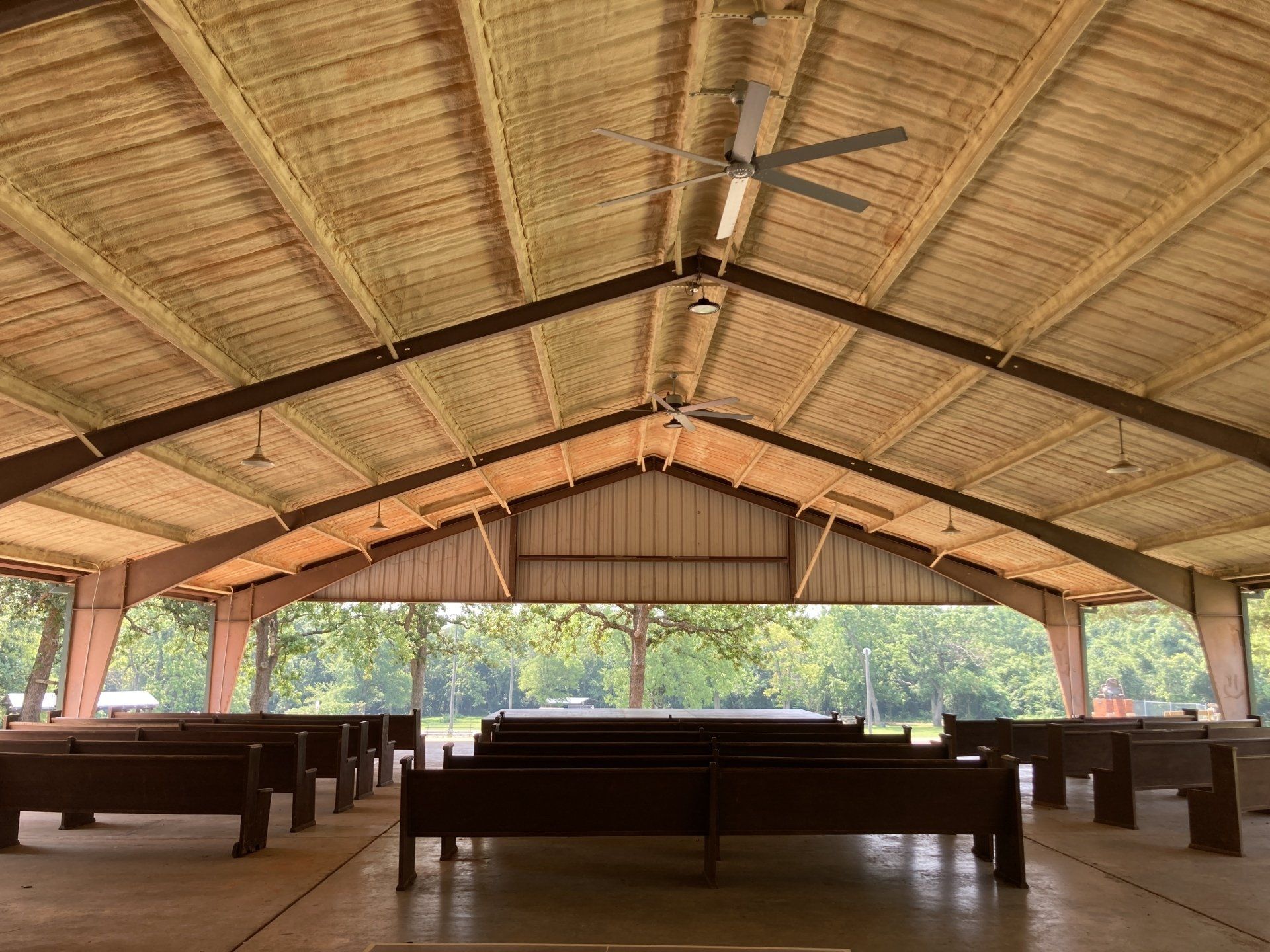 Inside view of an open-air pavilion with brown pews, a metal roof, and a view of trees through the open sides.