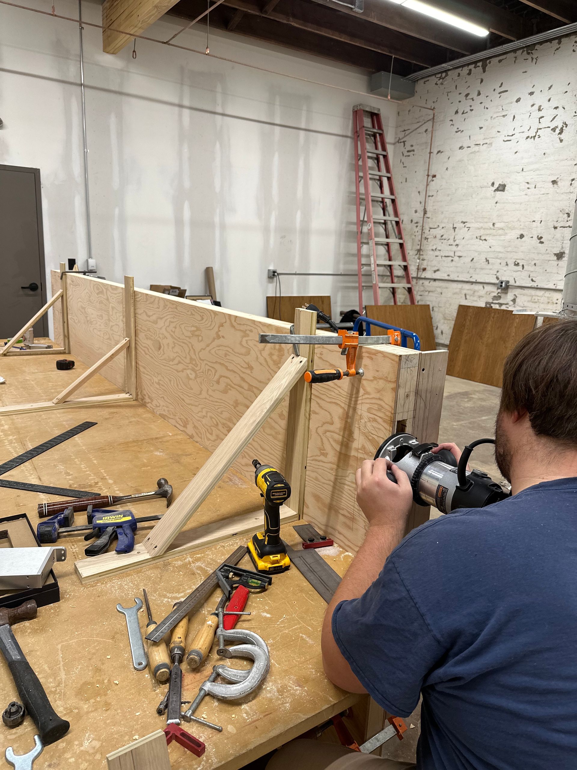 Man using a router on a wooden structure in a workshop, surrounded by tools and materials.